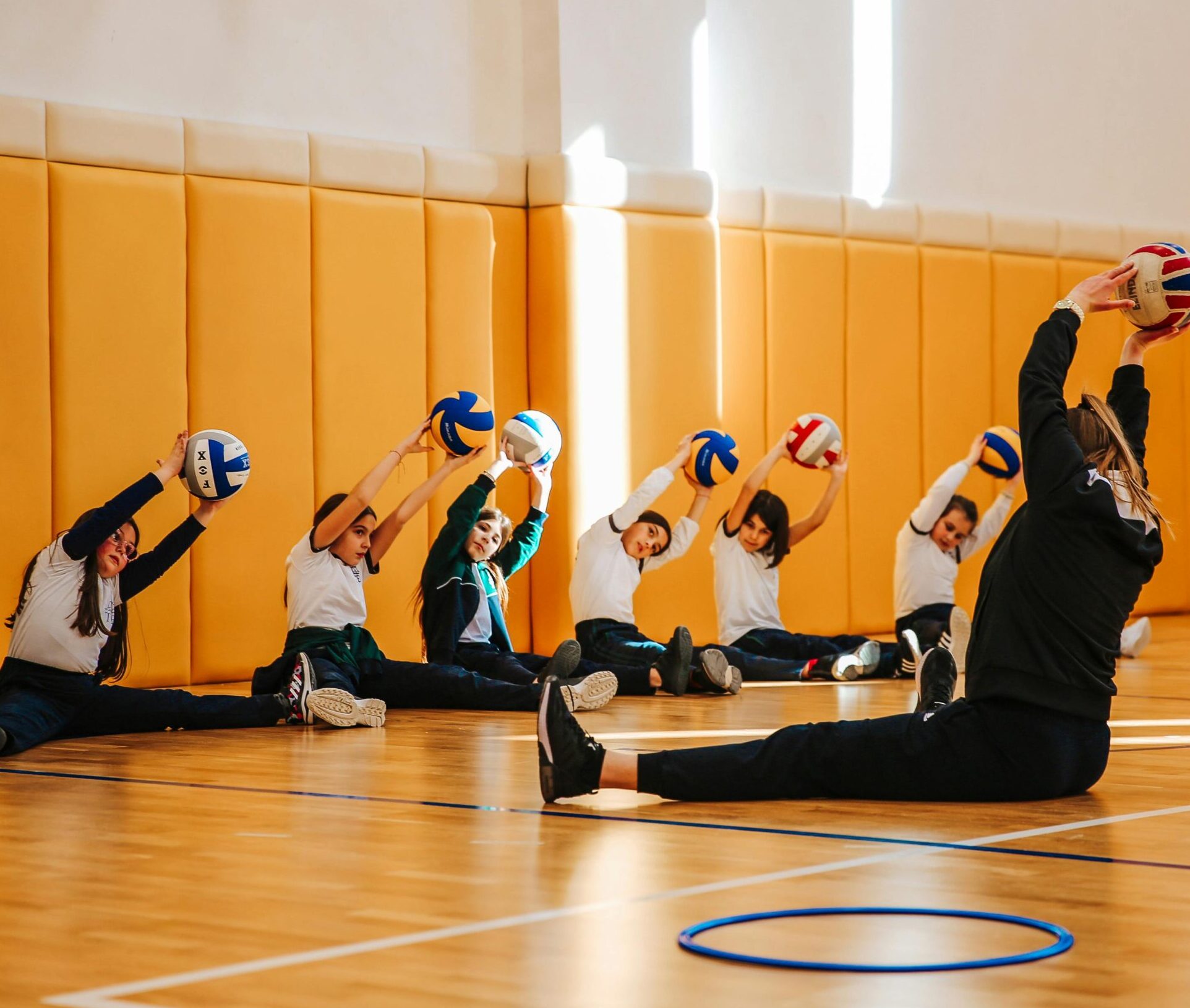 Group of children practicing volleyball stretches in a spacious indoor gym with a coach.