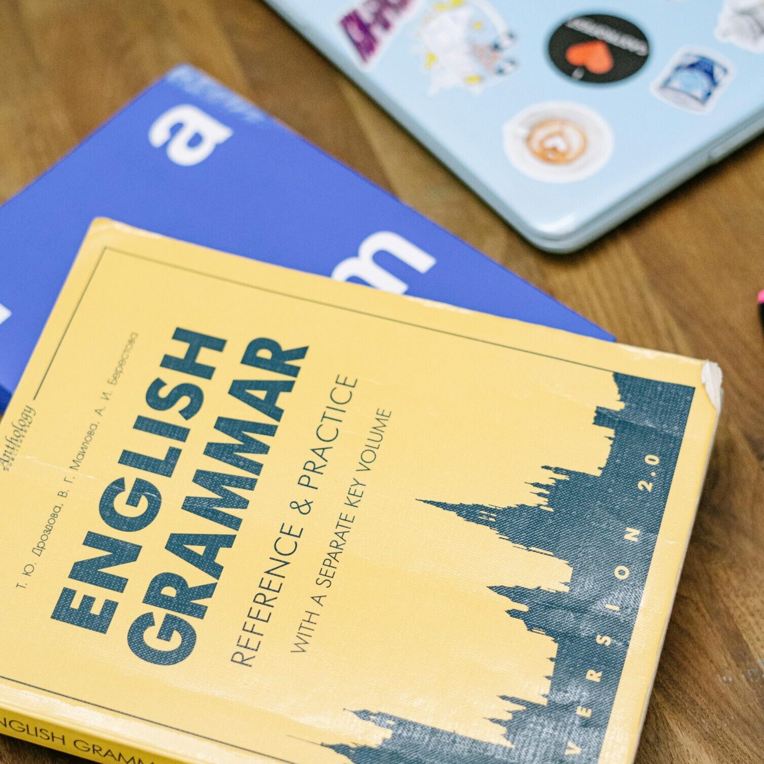 High-angle view of an English grammar book and a laptop with stickers on a wooden desk.